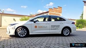 Biała Toyota Corolla z logo firmy Sunhelp i napisem 'Fotowoltaika, Pompy ciepła' zaparkowana przed budynkiem. Widoczne logo Visualpro w prawym dolnym rogu.