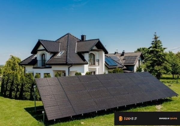 Instalacja fotowoltaiczna na gruncie przed nowoczesnym domem z panelami na dachu, widok z niskiej perspektywy, błękitne niebo.