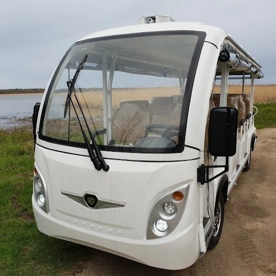 Biały, otwarty elektryczny bus turystyczny z widocznymi siedzeniami, stojący na tle jeziora i trzcin.