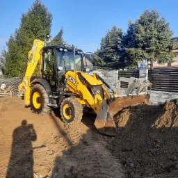 Żółta koparko-ładowarka JCB z łyżką nabierającą ziemię na placu budowy, w tle widoczne drzewa iglaste i ogrodzenie posesji, cień fotografa na pierwszym planie.