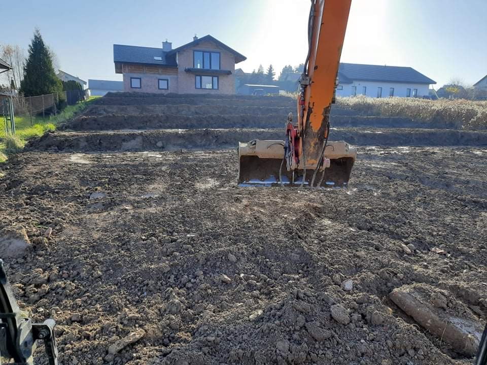 Pomarańczowa koparka w trakcie prac ziemnych na nierównym terenie pod budowę domu, widoczne stopnie w ziemi i dom w tle.