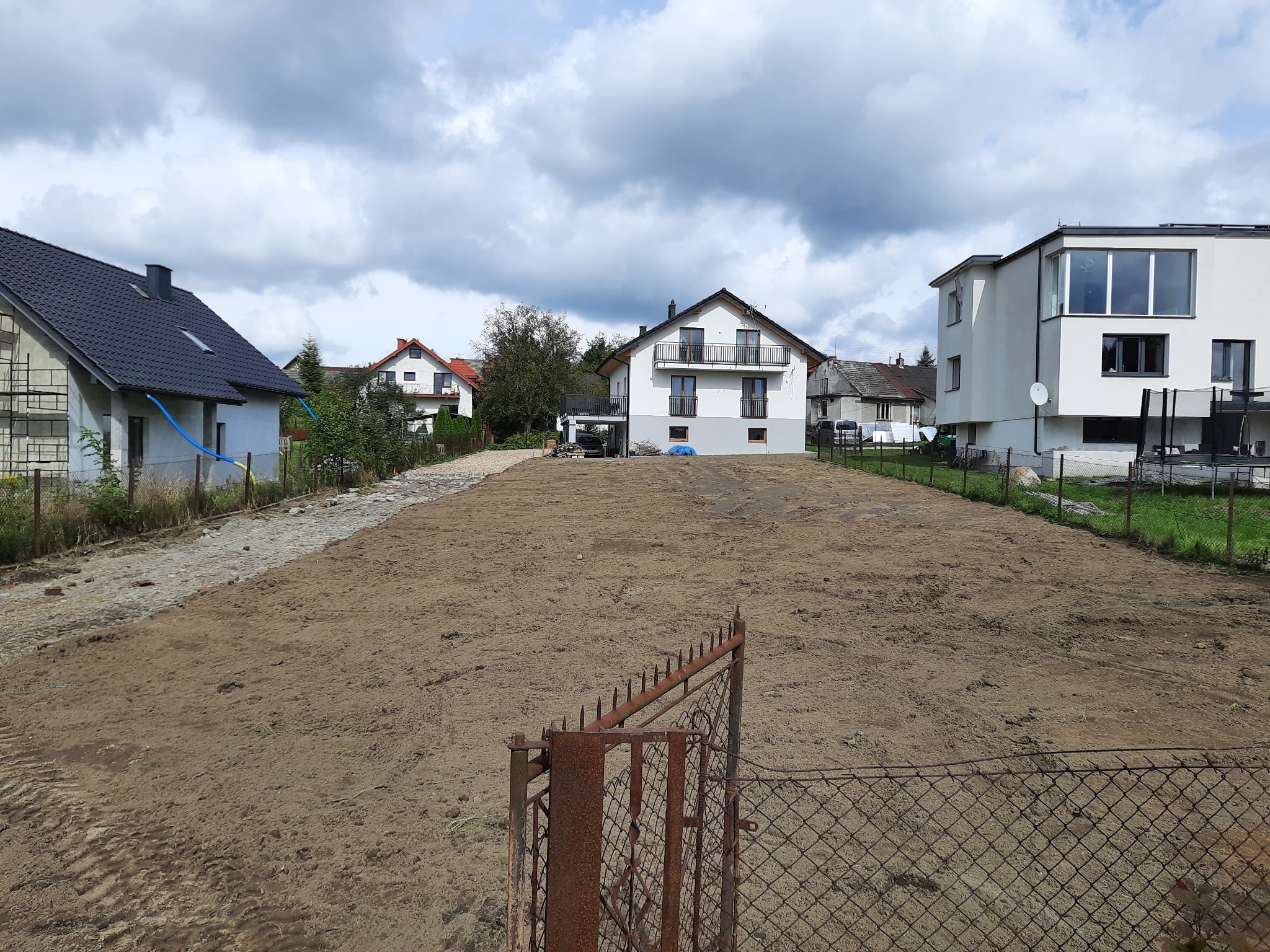 Przygotowany, wyrównany teren pod budowę z widocznymi domami w tle, ogrodzony zardzewiałą siatką i bramą.