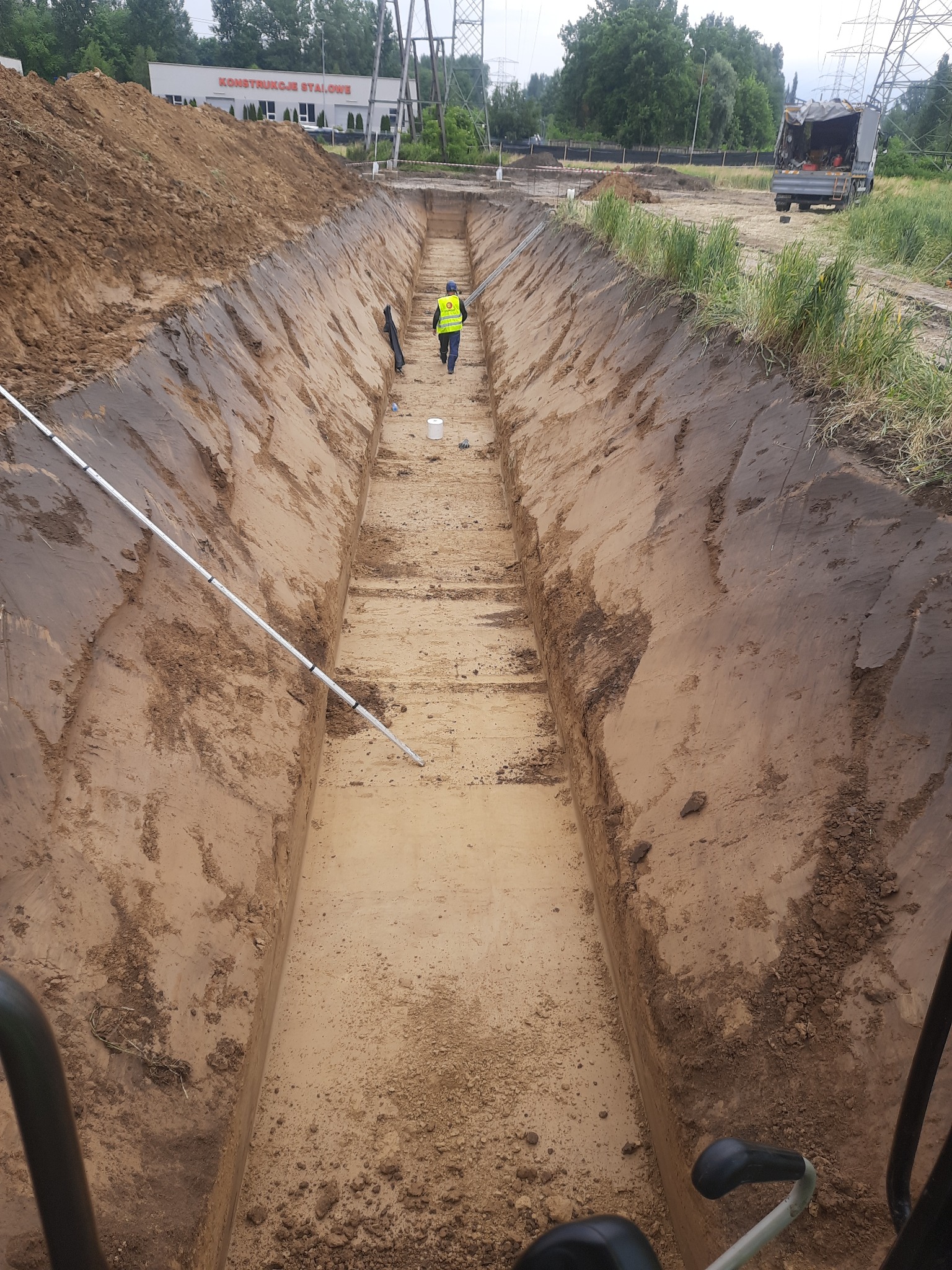 Długi, wąski wykop w ziemi z pracownikiem w kamizelce odblaskowej w oddali, widok z góry z kabiny maszyny, obok hałda ziemi i budynek 'Konstrukcje Stalowe'.