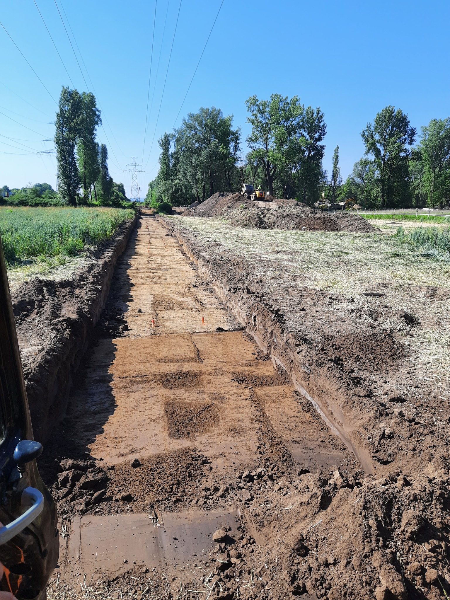 Długi wykop w ziemi z widocznymi śladami po maszynach, pomarańczowe znaczniki wzdłuż wykopu, w tle koparki i zwały ziemi, słupy wysokiego napięcia na horyzoncie.