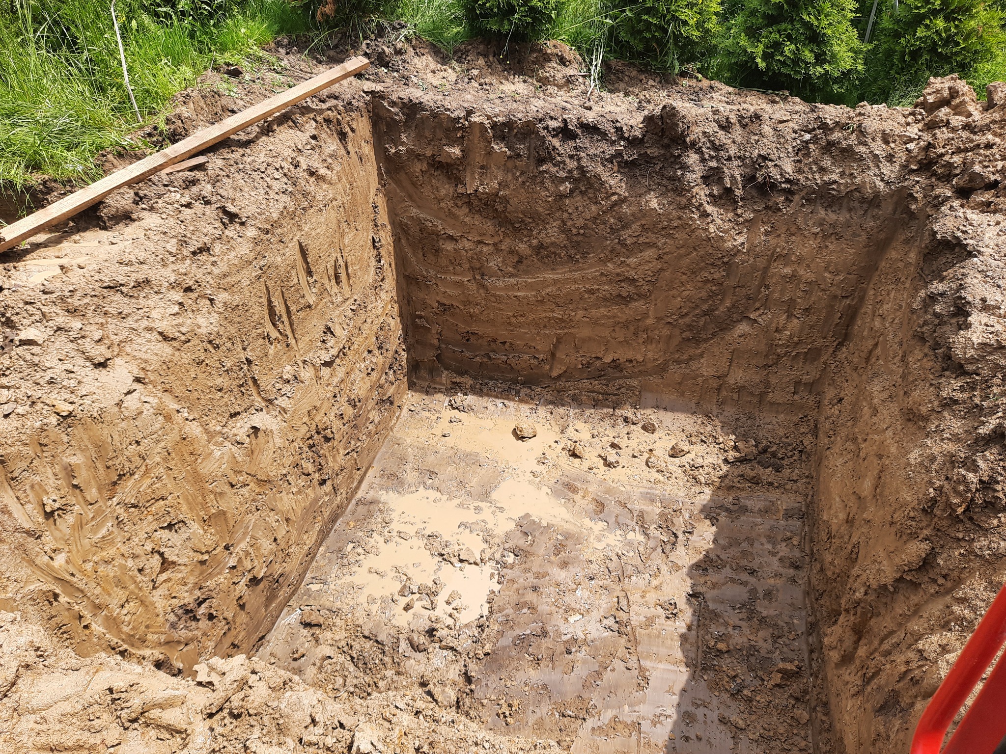 Głęboki, kwadratowy wykop w ziemi, częściowo wypełniony wodą i błotem, z deską leżącą na krawędzi i zielenią w tle.