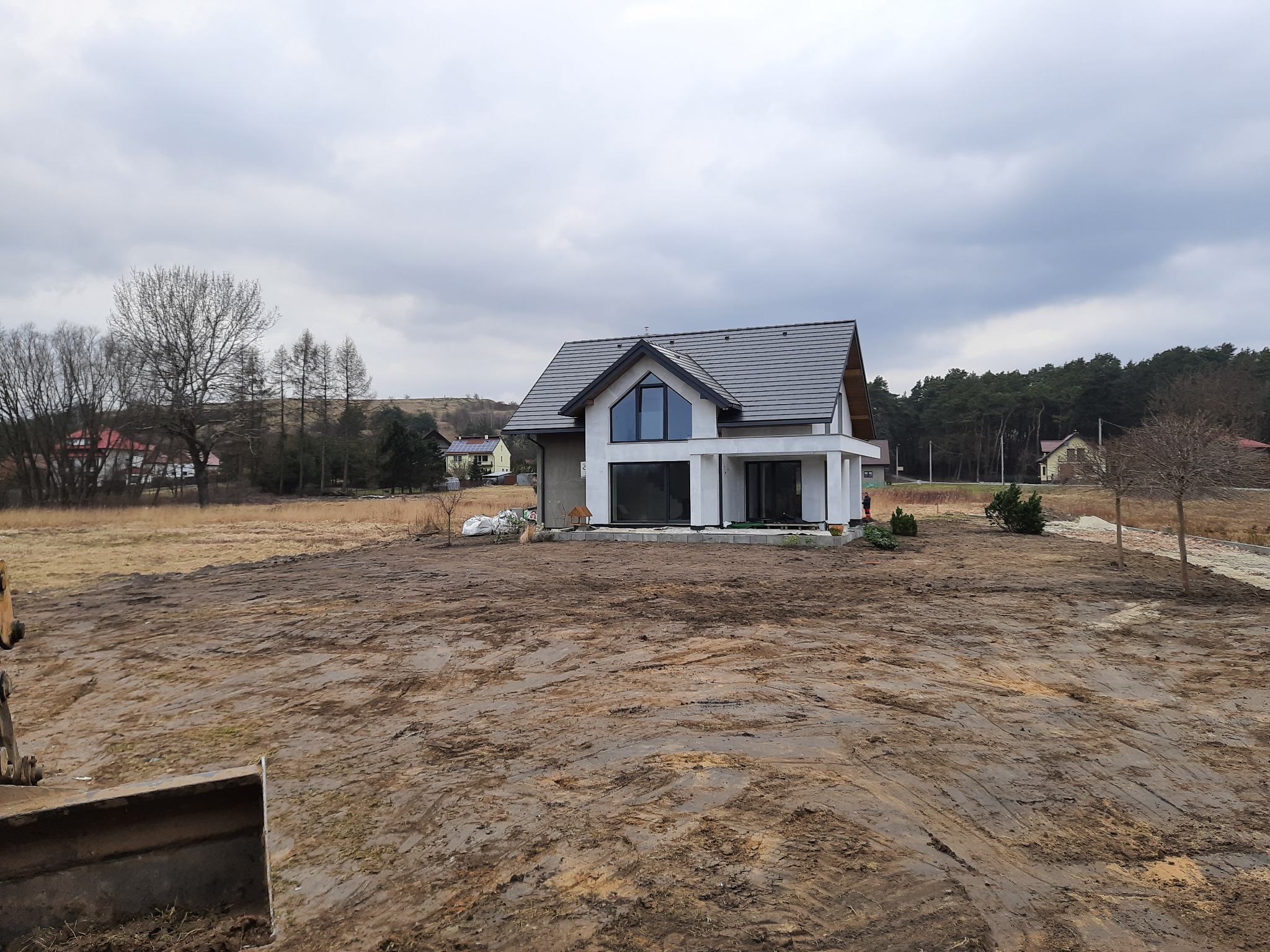 Nowoczesny dom jednorodzinny w trakcie budowy, widok na wyrównany teren działki z widocznymi śladami pracy maszyn budowlanych, pochmurne niebo.
