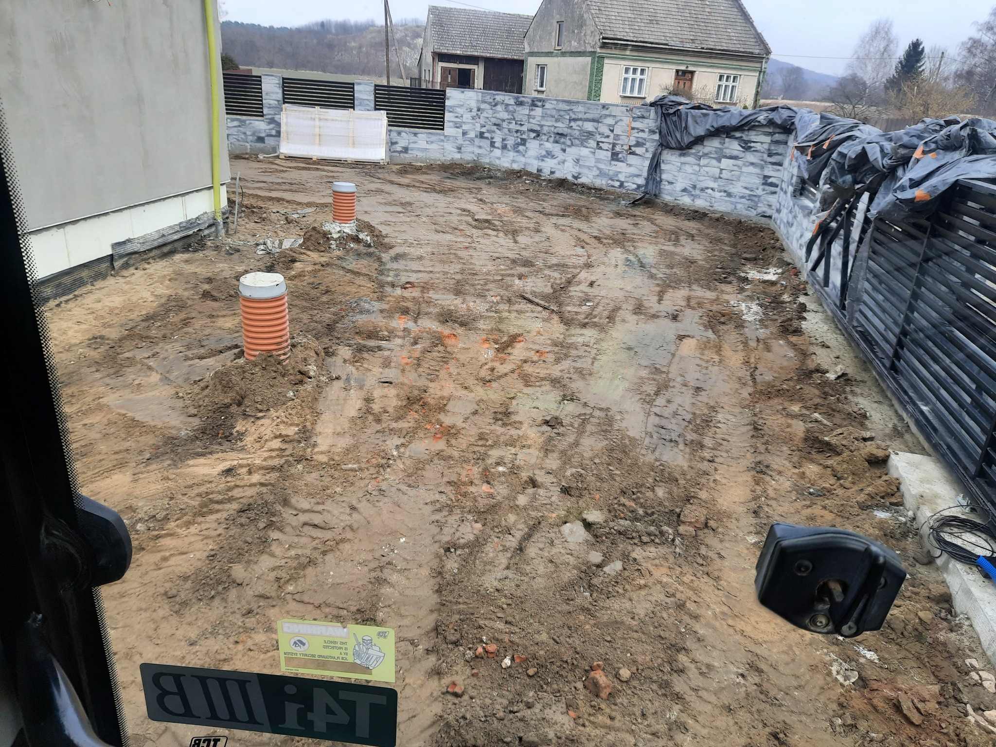 Przygotowany teren pod budowę domu, widoczne rury kanalizacyjne, świeżo wyrównana ziemia z śladami opon, ogrodzenie z betonowych bloczków i metalowych przęseł w tle, widok z kabiny maszyny budowlanej.