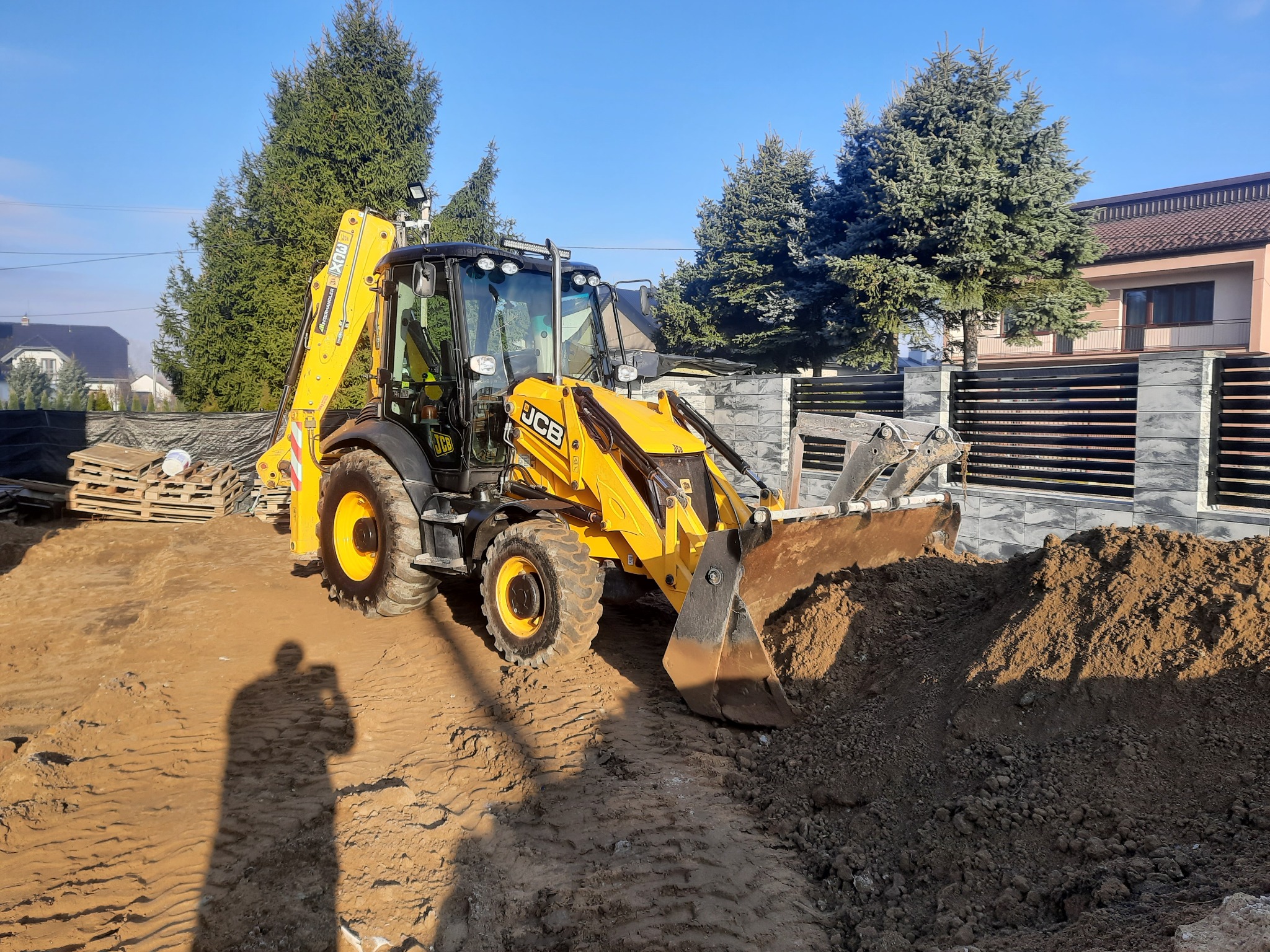 Żółta koparko-ładowarka JCB z łyżką nabierającą ziemię na placu budowy, w tle widoczne drzewa iglaste i ogrodzenie posesji, cień fotografa na pierwszym planie.
