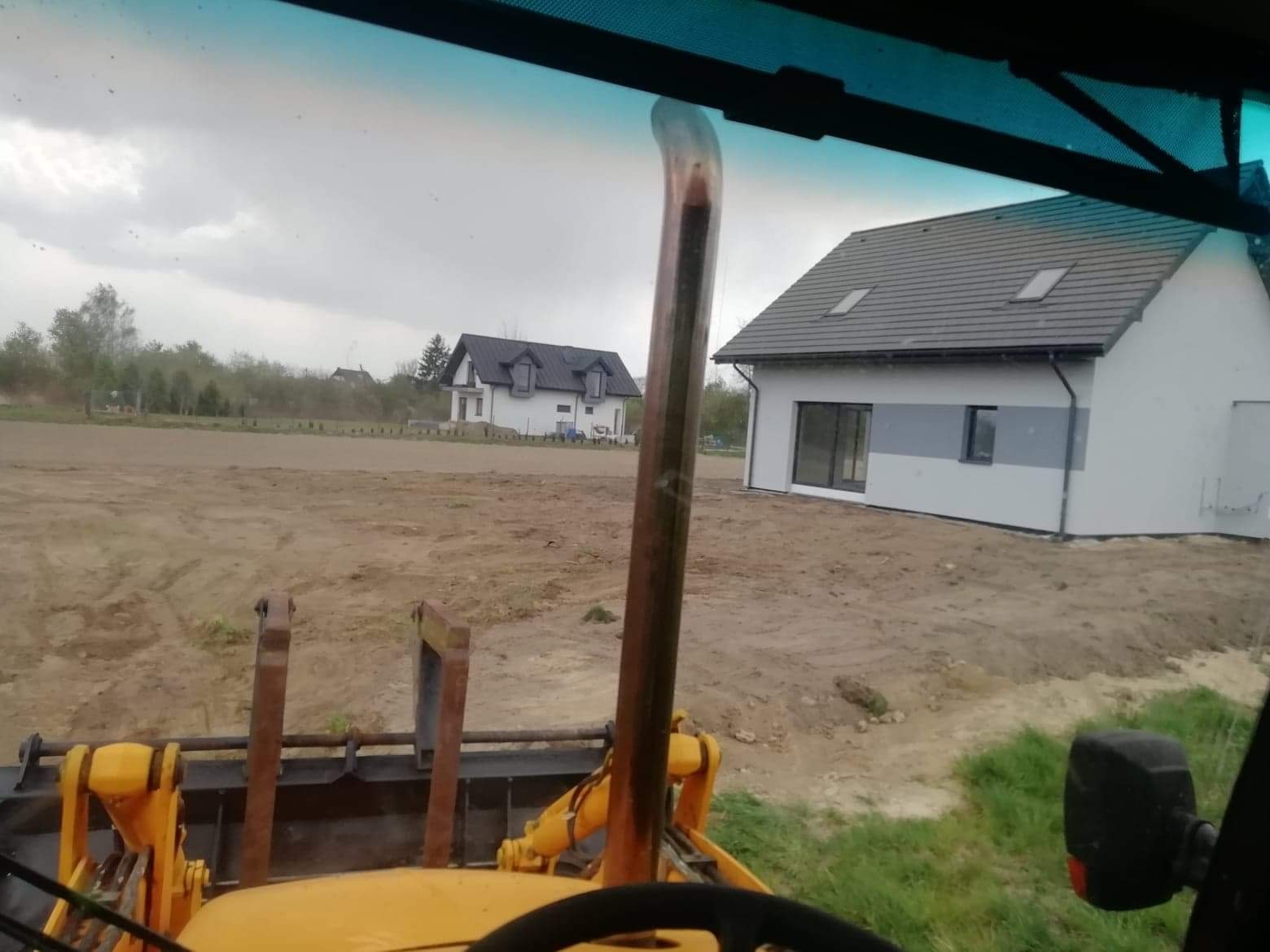 Widok z kabiny koparko-ładowarki na wyrównany teren budowy z dwoma nowymi domami w tle, pochmurne niebo.