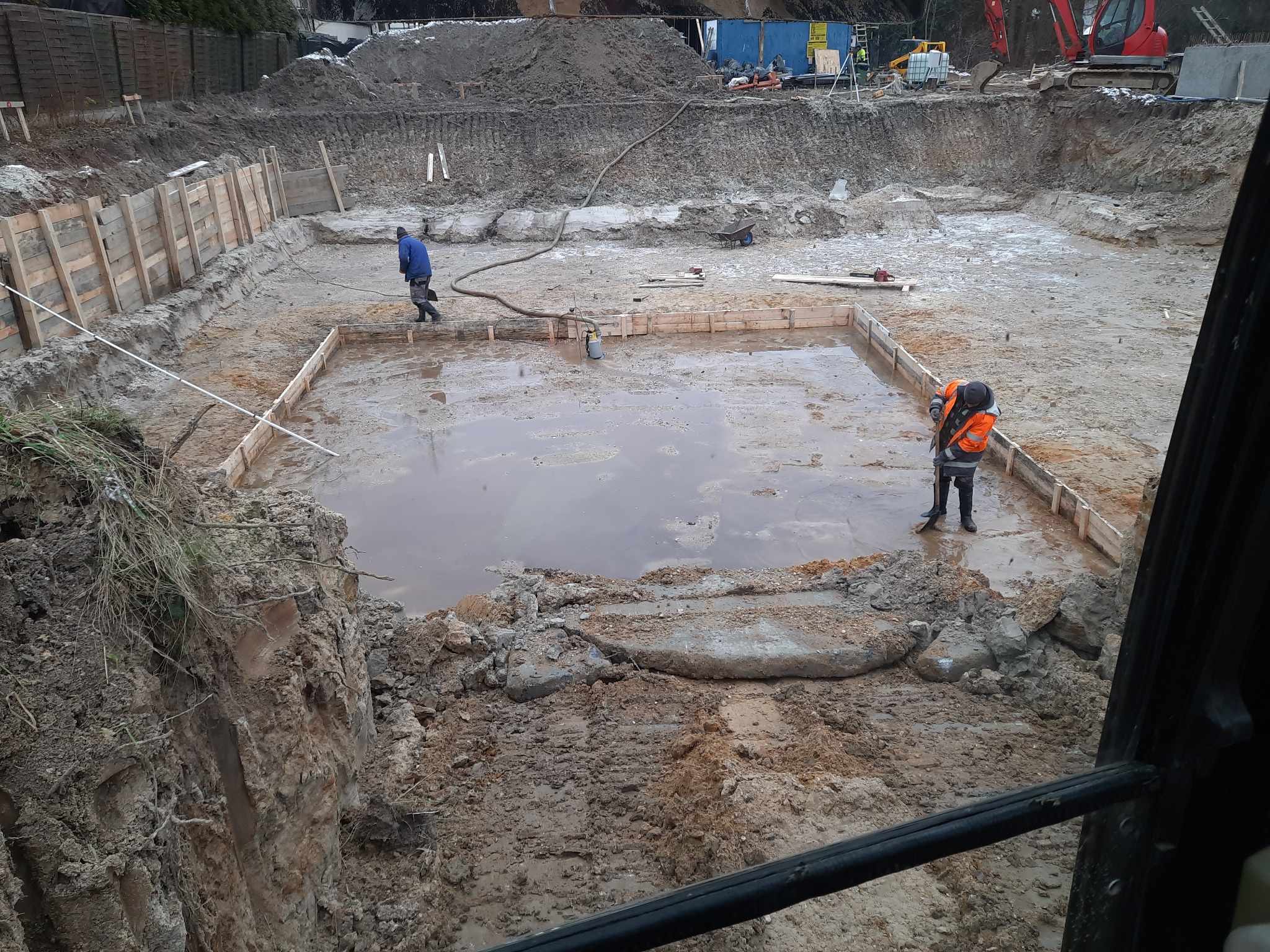 Wykop pod fundamenty z drewnianym szalunkiem, częściowo zalany wodą, dwóch pracowników przy pracy, w tle koparka i hałda ziemi.