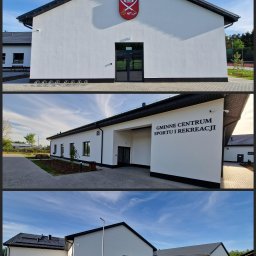 TaFFa-Dawid Wnętrzak - Nowoczesny budynek Gminnego Centrum Sportu i Rekreacji z białą elewacją, czarnym dachem i herbem gminy nad wejściem, otoczony zagospodarowanym terenem z chodnikami i zielenią.