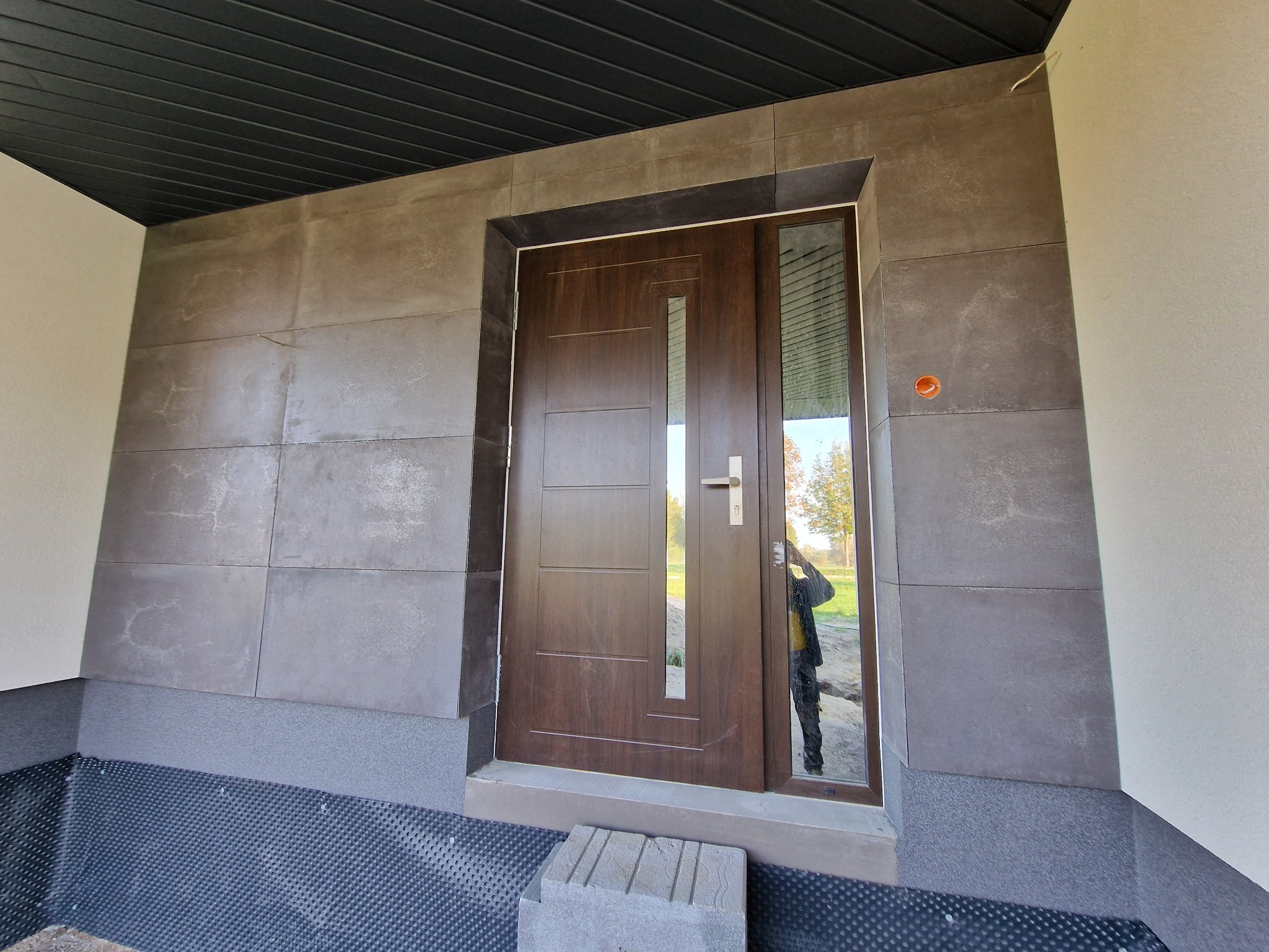 Wejście do domu z brązowymi drzwiami i elewacją wykończoną płytkami imitującymi kamień, widoczna perforowana membrana fundamentowa i odbicie fotografa w szybie drzwi.