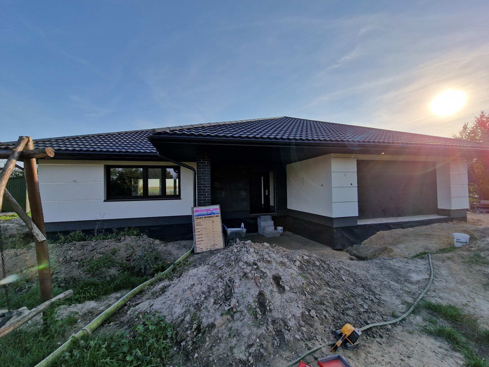 Nowoczesny dom jednorodzinny z ciemną elewacją garażu i białymi ścianami, widoczny plac budowy z hałdą ziemi i tablicą informacyjną przy wejściu.