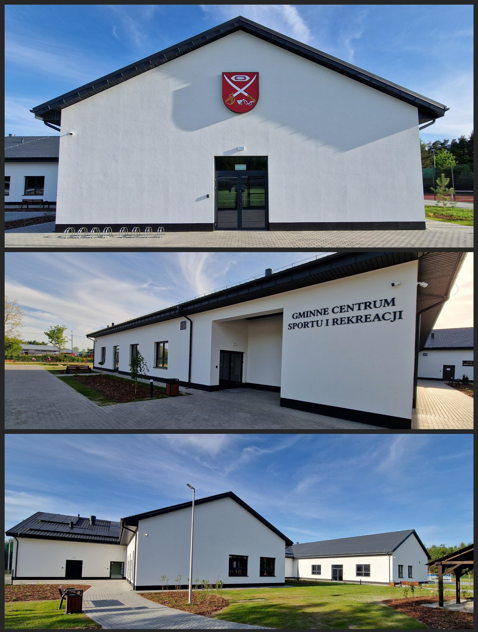 Nowoczesny budynek Gminnego Centrum Sportu i Rekreacji z białą elewacją, czarnym dachem i herbem gminy nad wejściem, otoczony zagospodarowanym terenem z chodnikami i zielenią.