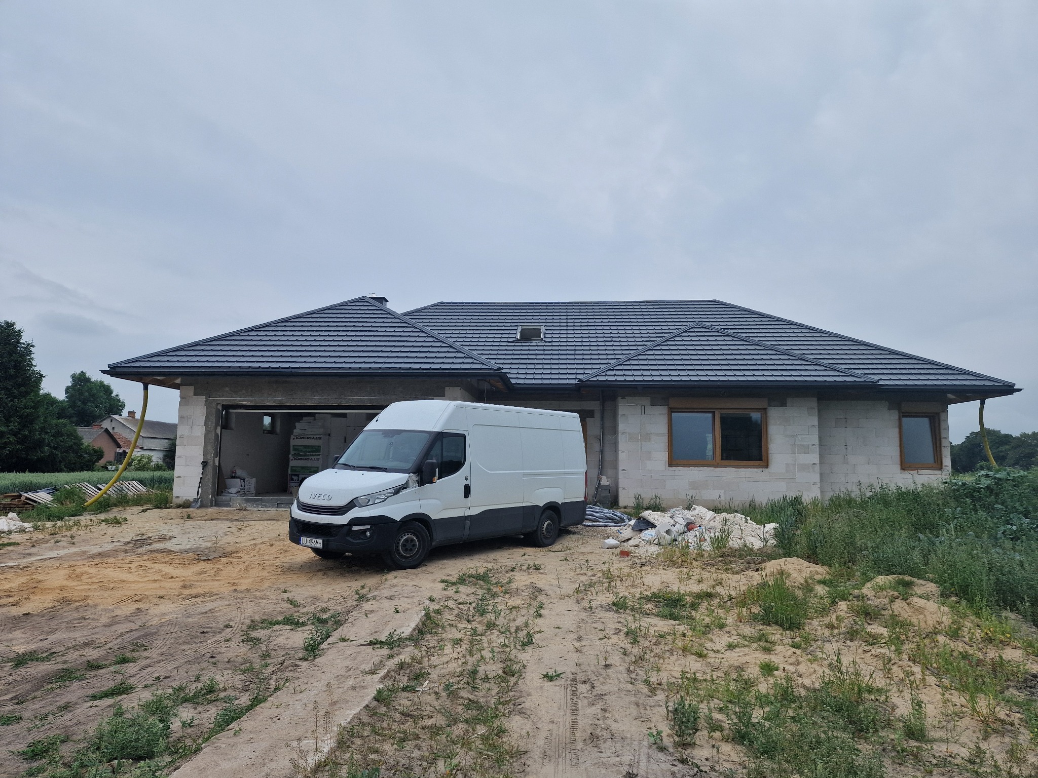 Surowy stan domu jednorodzinnego z szarą dachówką, biały van Iveco zaparkowany przed garażem, materiały budowlane składowane obok.