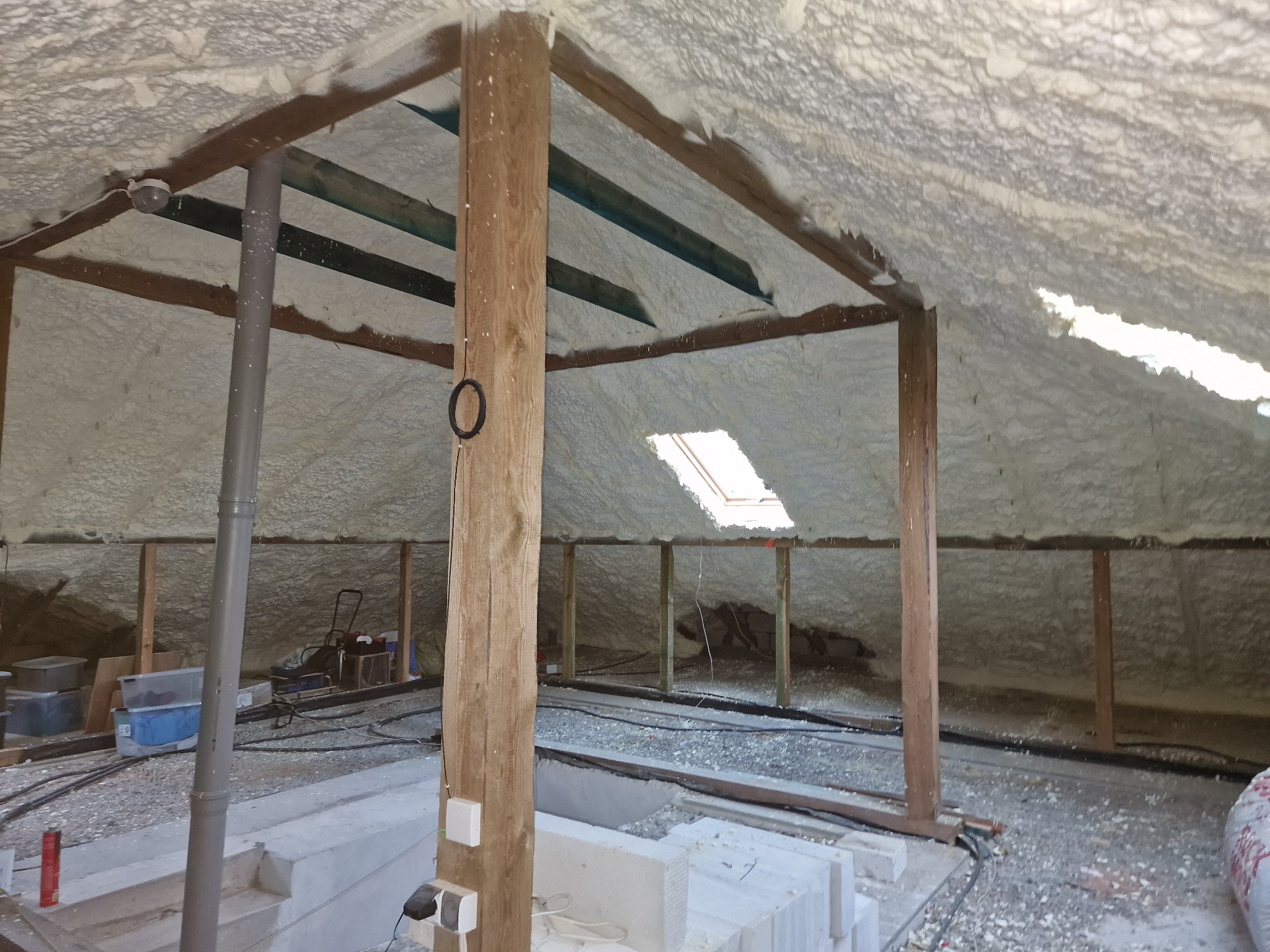 Poddasze ocieplone pianką poliuretanową, widoczne drewniane elementy konstrukcyjne, okno dachowe i instalacje elektryczne. Częściowo wykończona podłoga z widocznymi bloczkami betonu komórkowego.