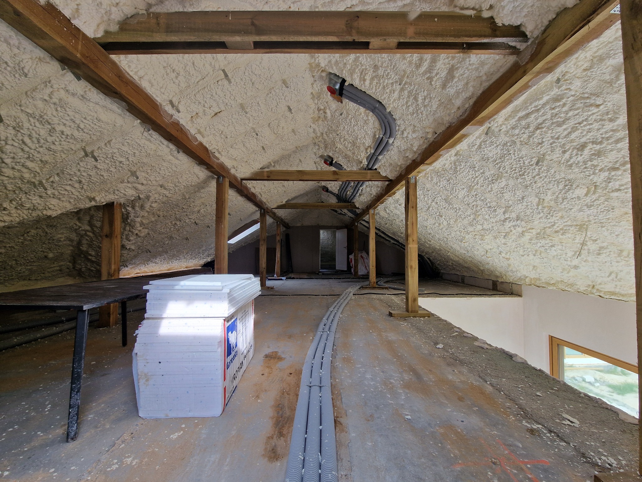 Poddasze z drewnianą konstrukcją pokryte pianką izolacyjną, widoczne szare rury instalacyjne, stos płyt izolacyjnych i fragment okna dachowego.