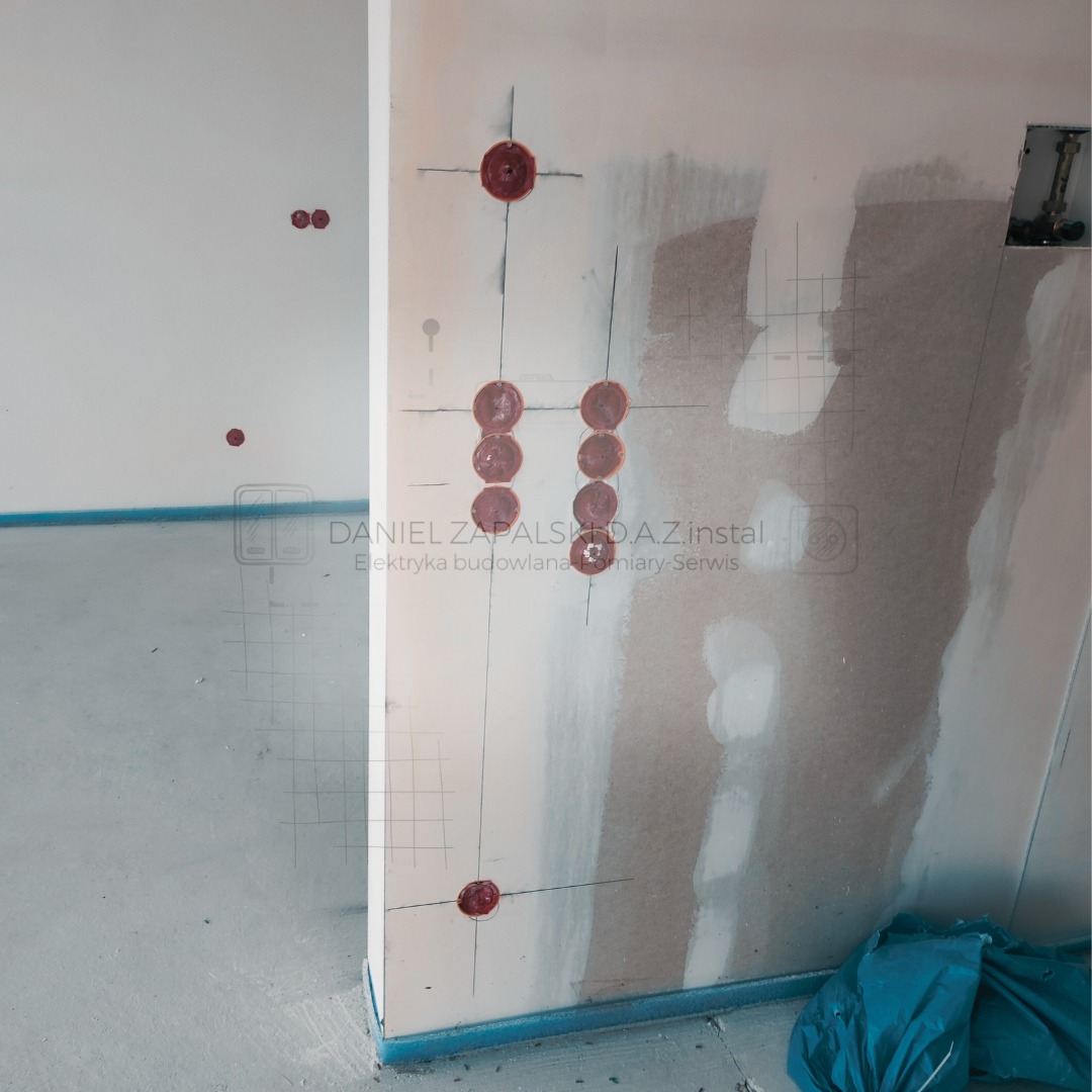 Przygotowanie do montażu gniazdek elektrycznych w nowym budynku, widoczne wywiercone otwory w ścianie i zaznaczone punkty, częściowo pokryte gipsem.