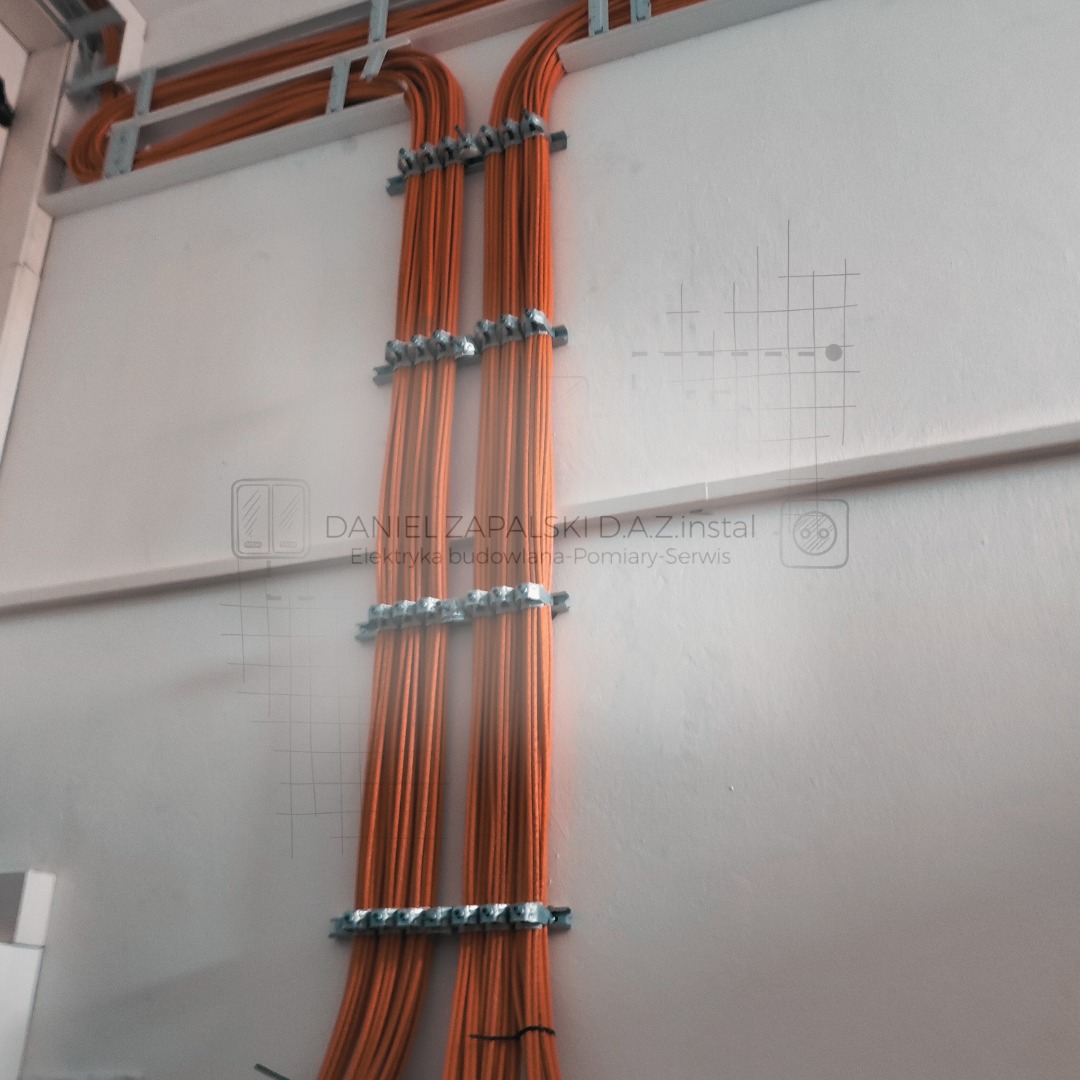 Dwa pionowe pęki pomarańczowych kabli elektrycznych, przymocowane metalowymi uchwytami do jasnej ściany, z widocznymi elementami instalacji pod sufitem.