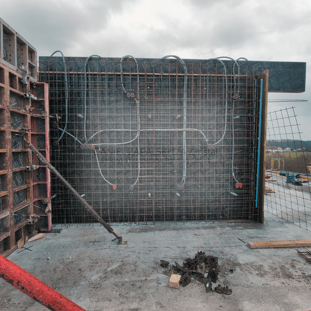 Szare przewody elektryczne ułożone wewnątrz metalowej siatki zbrojeniowej, przygotowane do zalania betonem w szalunku na budowie, widoczne elementy mocujące i fragment krajobrazu w tle.