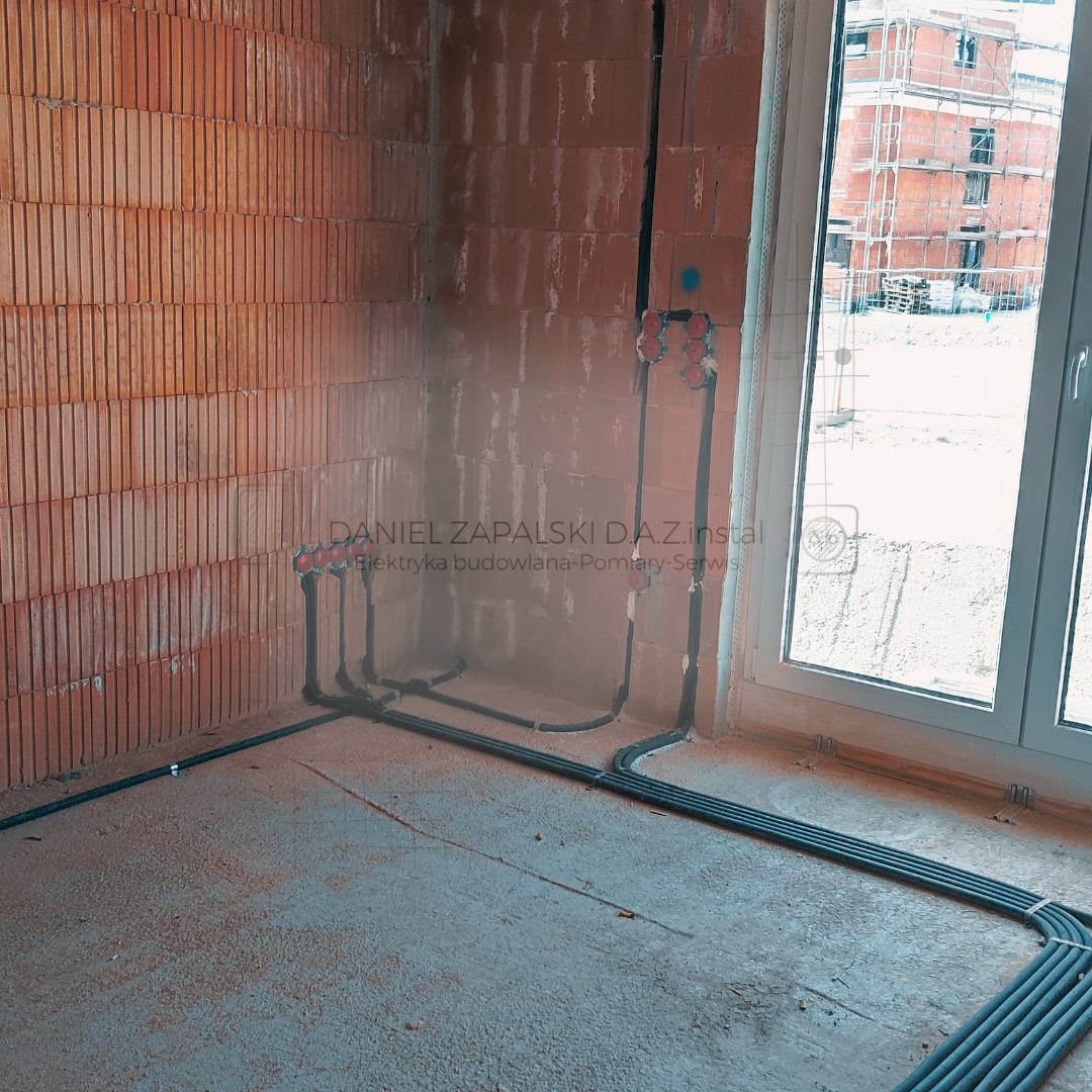 Surowe wnętrze z ceglanymi ścianami, widoczne rury instalacji elektrycznej prowadzone po ścianach i podłodze, z puszkami pod gniazdka, okno z widokiem na budynek w budowie.