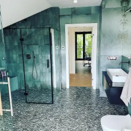 Nowoczesna łazienka w odcieniach szarości i zieleni z kabiną prysznicową typu walk-in, umywalką nablatową i oryginalną mozaiką na podłodze, widok na sąsiednie pomieszczenie z drewnianą podłogą...