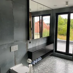 Nowoczesna łazienka z szarymi płytkami imitującymi beton na ścianach, podwieszaną toaletą, czarną szafką z umywalką i dużym lustrem, widok na zewnątrz przez okno.