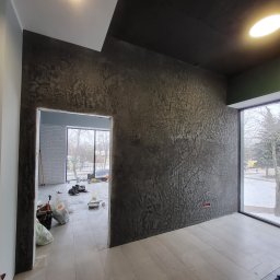 Piotr Gągolewski - Pokój w trakcie prac, ściana pokryta dekoracyjną, ciemną strukturą, widoczne worki z materiałami i narzędzia, okno z widokiem na zimowy krajobraz.