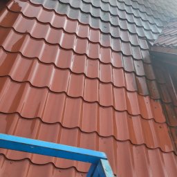 Piotr Gągolewski - Czerwona blachodachówka po deszczu, widoczna z bliska z fragmentem niebieskiego rusztowania, na części dachu widoczne ślady mchu.