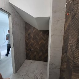 Piotr Gągolewski - Wnętrze łazienki w trakcie prac wykończeniowych: ściany z płytek imitujących marmur, fragment ściany wyłożony drewnopodobną mozaiką w jodełkę, widoczne przewody instalacji, w tle sylwetka osoby...