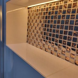 Piotr Gągolewski - Narożna wnęka z półką, wyłożona mozaiką w odcieniach brązu i szarości, oświetlona listwą LED od góry.