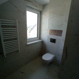 Piotr Gągolewski - Wykończona łazienka z białymi płytkami imitującymi marmur ze złotymi żyłami, wisząca toaleta z czarnym przyciskiem spłukiwania, dekoracyjna mozaika nad toaletą, biały grzejnik drabinkowy, okno...