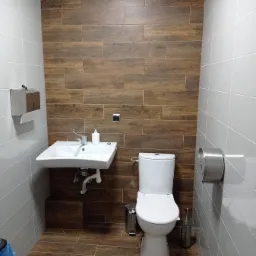 Nowoczesna łazienka z płytkami imitującymi drewno na ścianie i podłodze, biała umywalka i toaleta, metalowe akcesoria.