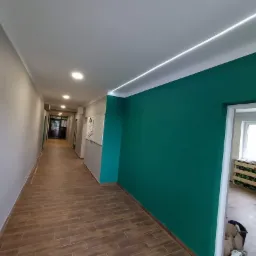 Długi korytarz po malowaniu: ściany w odcieniach szarości i butelkowej zieleni, drewnopodobna podłoga, oświetlenie liniowe LED w suficie oraz punktowe, widok na pomieszczenie z oknem.