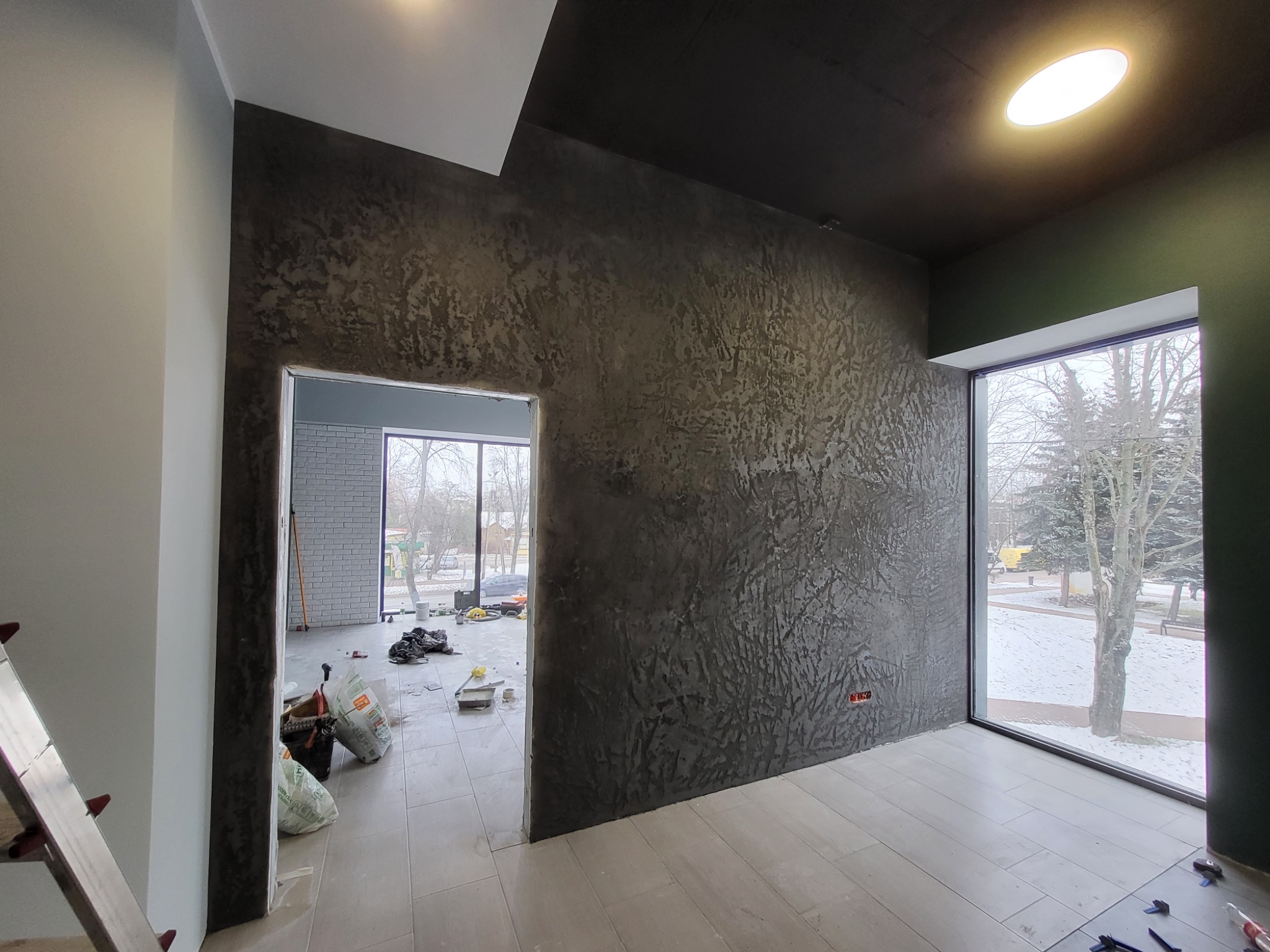 Pokój w trakcie prac, ściana pokryta dekoracyjną, ciemną strukturą, widoczne worki z materiałami i narzędzia, okno z widokiem na zimowy krajobraz.