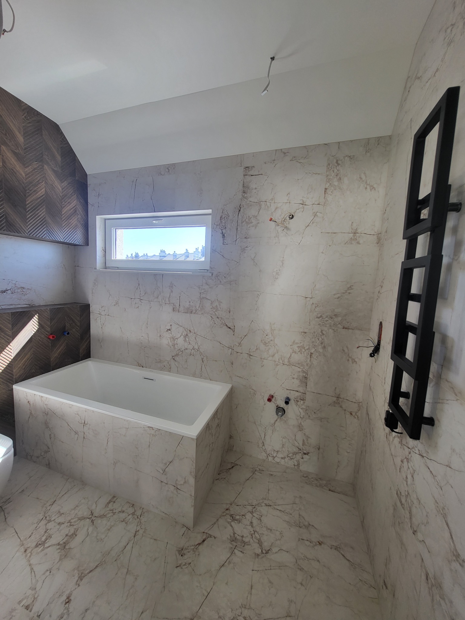 Nowoczesna łazienka z wanną obłożoną marmurowymi płytkami, dekoracyjną ścianą z drewnianą mozaiką w jodełkę i czarnym grzejnikiem drabinkowym.