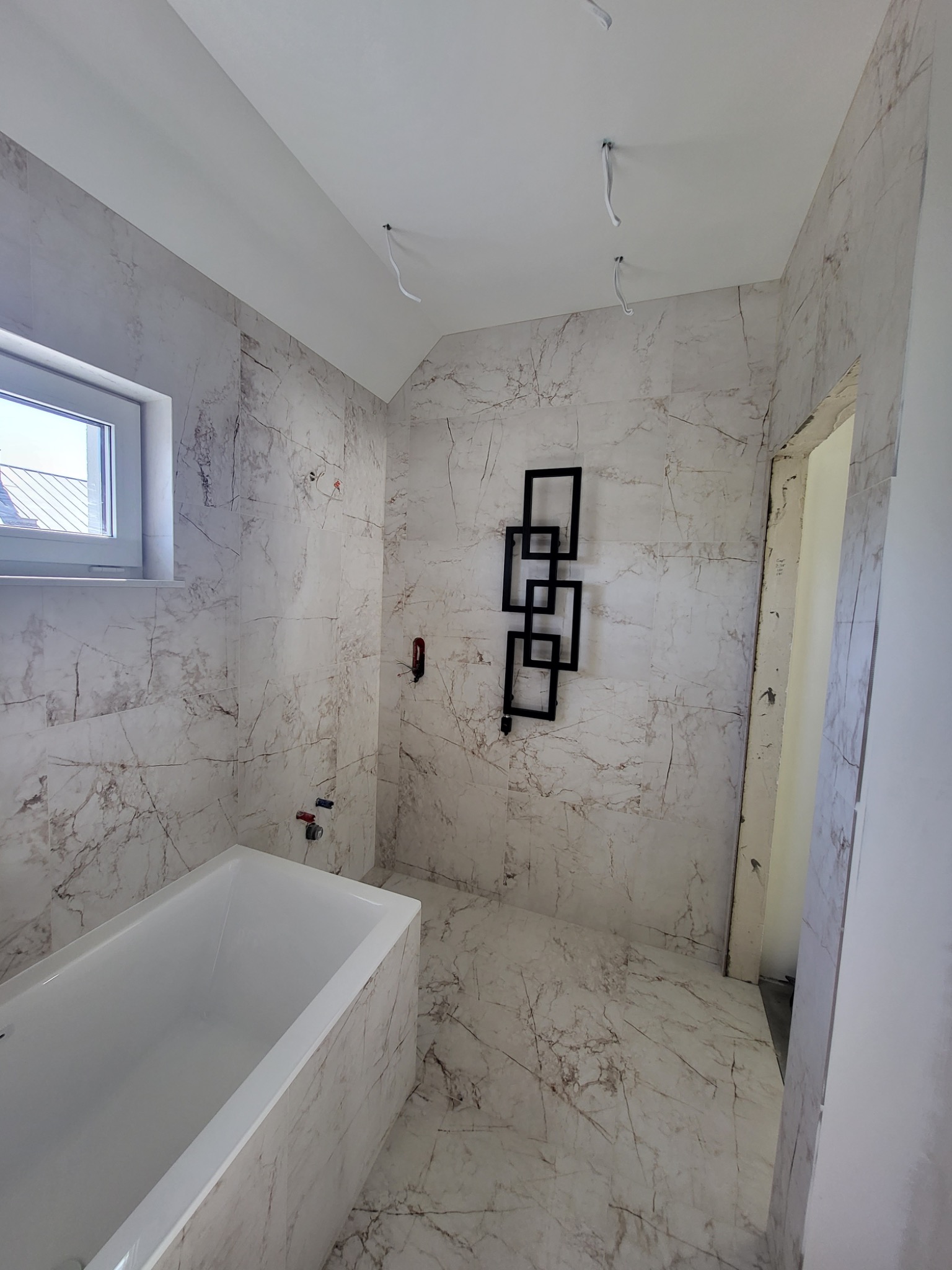 Wnętrze łazienki w trakcie remontu z wanną, płytkami imitującymi marmur na ścianach i podłodze, oraz dekoracyjnym grzejnikiem w kształcie geometrycznych figur. Widoczne wystające kable elektryczne...