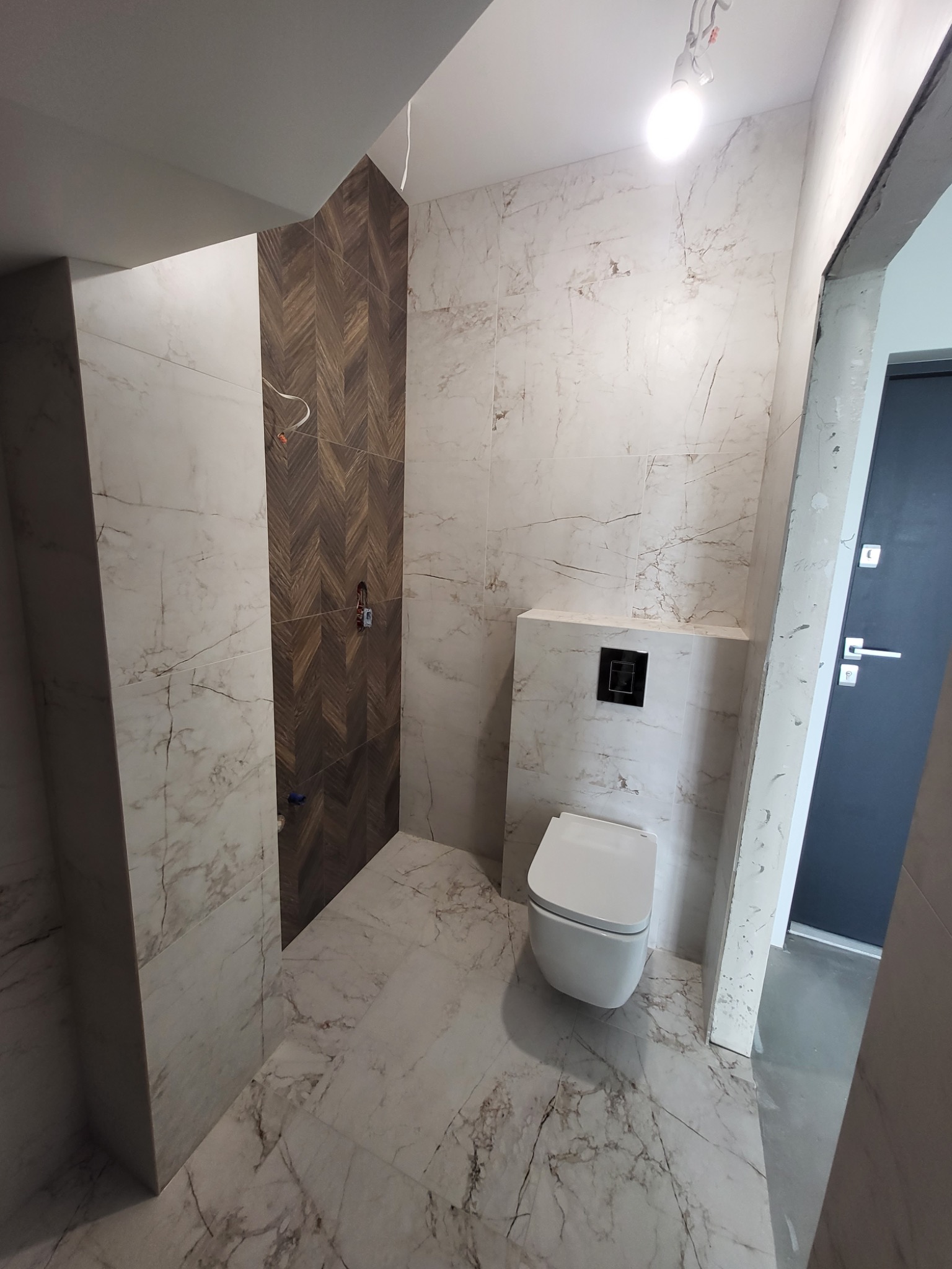 Nowoczesna łazienka w trakcie remontu: białe płytki imitujące marmur na ścianach i podłodze, kontrastująca ściana z drewnopodobnych płytek ułożonych w jodełkę, wisząca toaleta, widoczne...