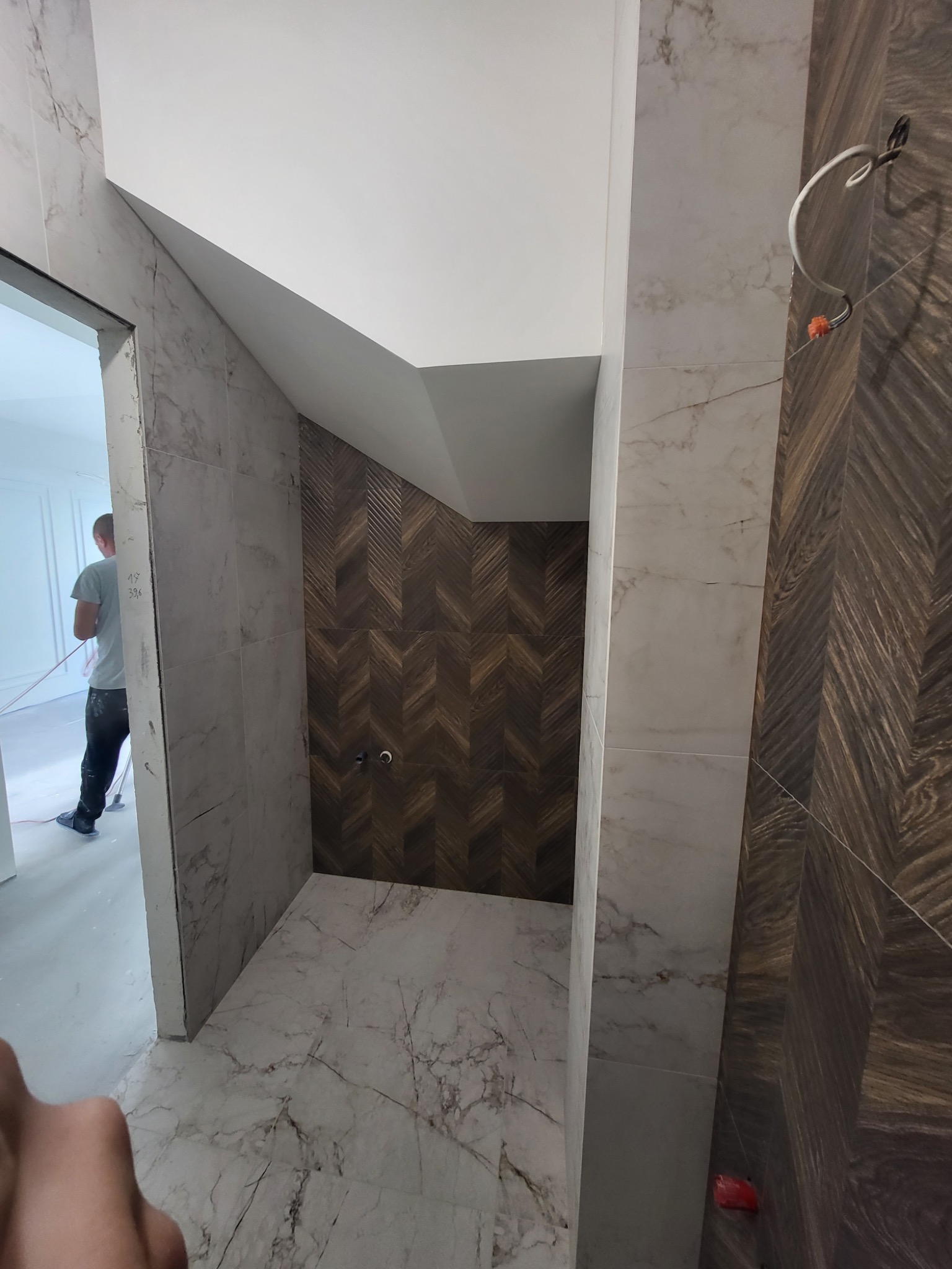 Wnętrze łazienki w trakcie prac wykończeniowych: ściany z płytek imitujących marmur, fragment ściany wyłożony drewnopodobną mozaiką w jodełkę, widoczne przewody instalacji, w tle sylwetka osoby...