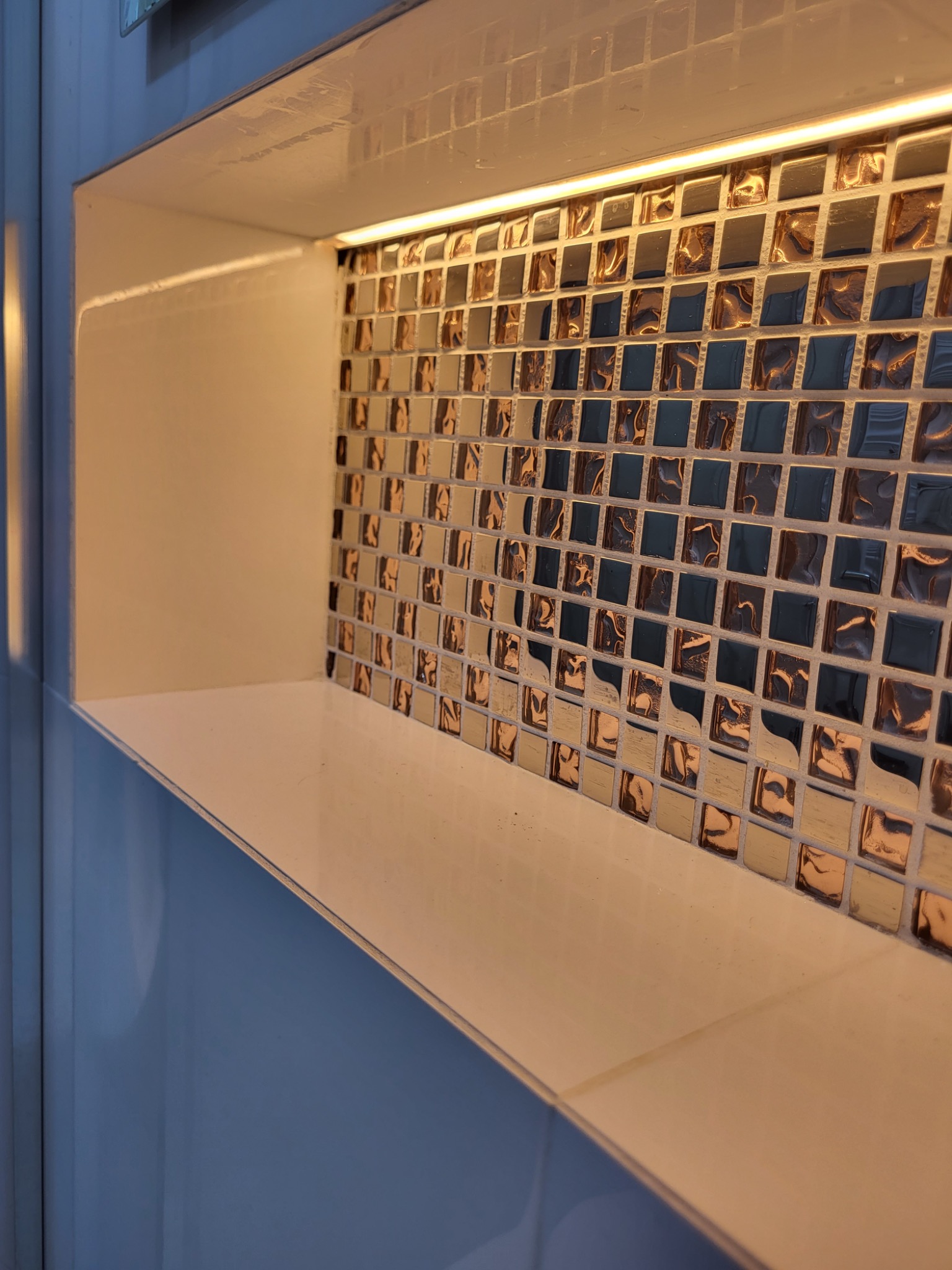 Narożna wnęka z półką, wyłożona mozaiką w odcieniach brązu i szarości, oświetlona listwą LED od góry.