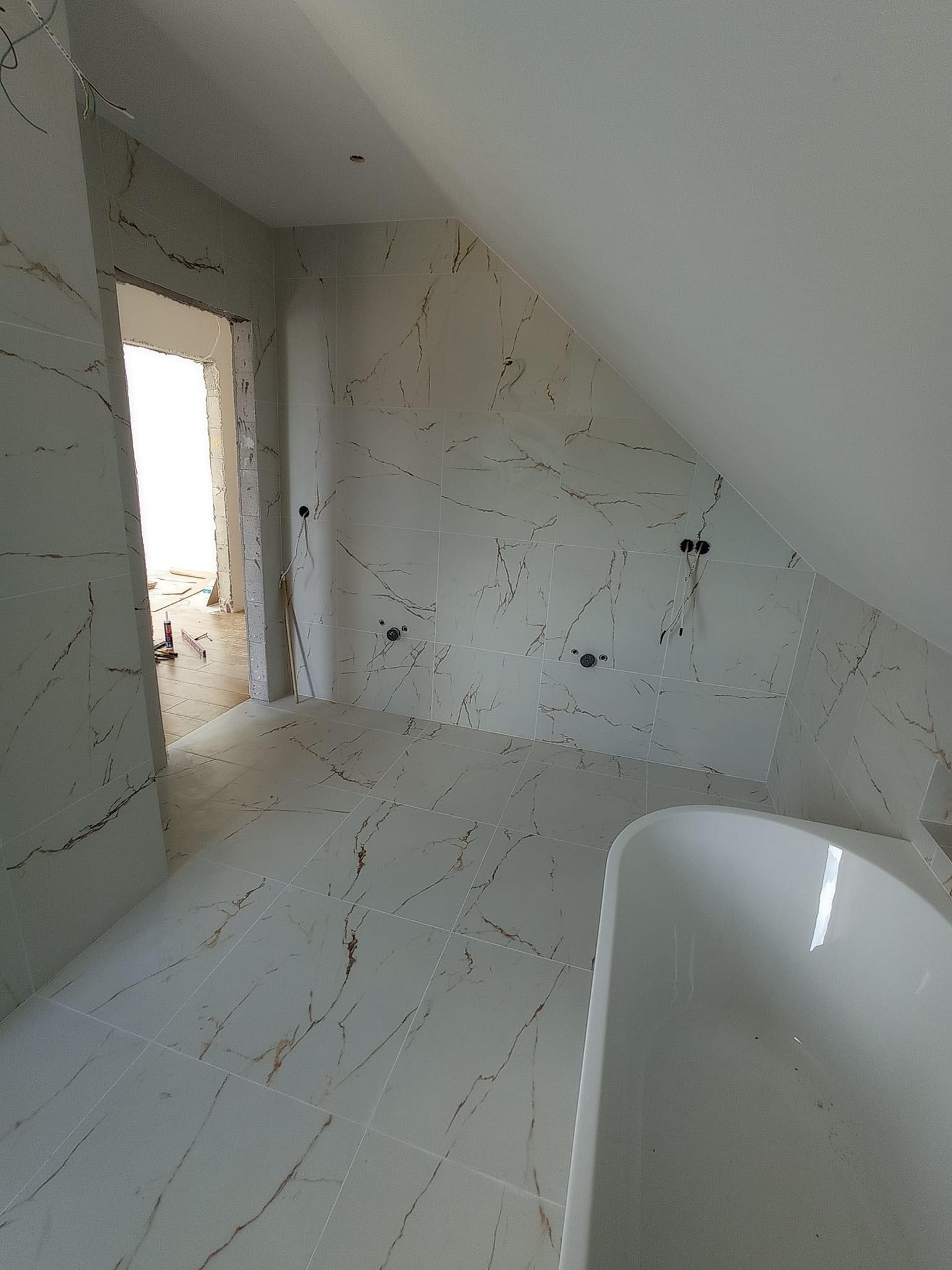 Białe płytki imitujące marmur na ścianach i podłodze łazienki na poddaszu, widoczne przygotowane otwory na instalacje, fragment wanny i otwarte drzwi do innego pomieszczenia w trakcie prac.