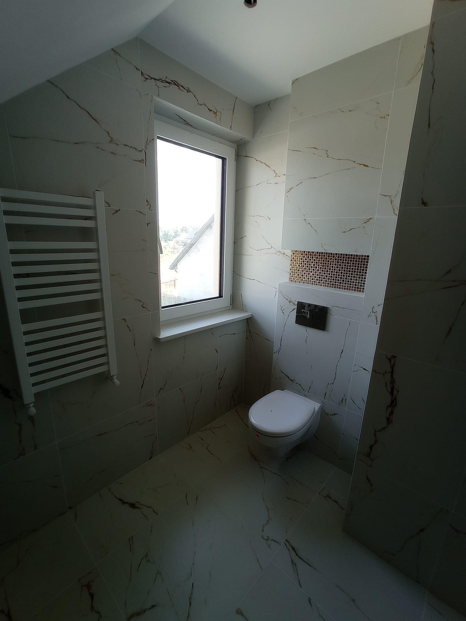 Wykończona łazienka z białymi płytkami imitującymi marmur ze złotymi żyłami, wisząca toaleta z czarnym przyciskiem spłukiwania, dekoracyjna mozaika nad toaletą, biały grzejnik drabinkowy, okno...