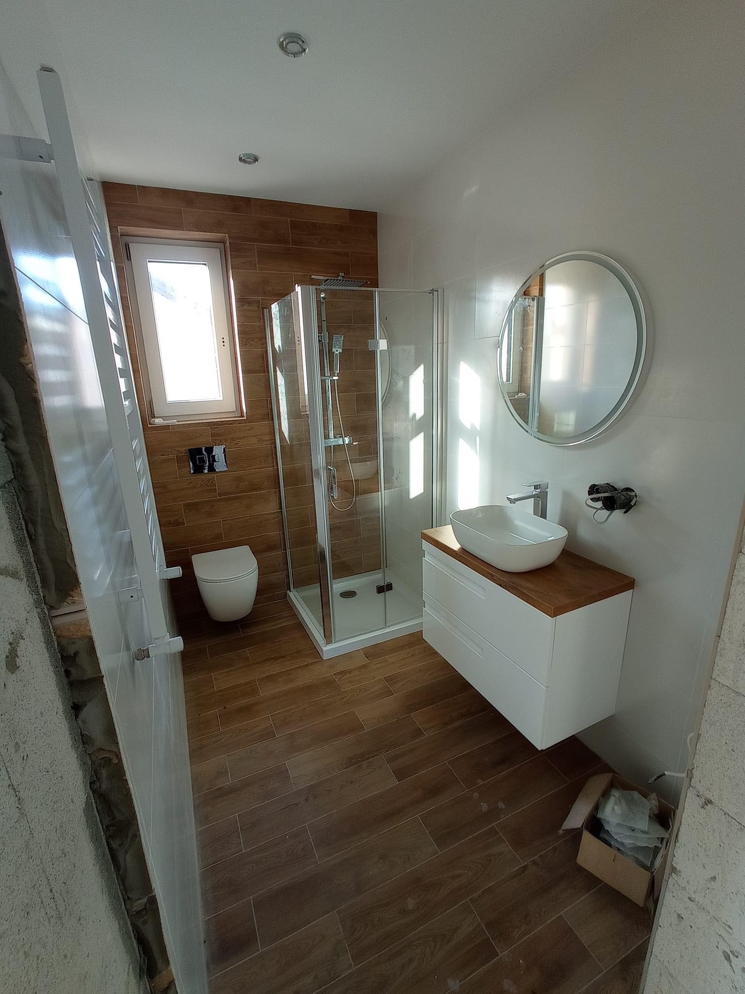 Nowoczesna łazienka w trakcie wykańczania: kabina prysznicowa z białymi ramami, umywalka nablatowa na białej szafce z blatem drewnopodobnym, okrągłe lustro, toaleta pod oknem, płytki imitujące...