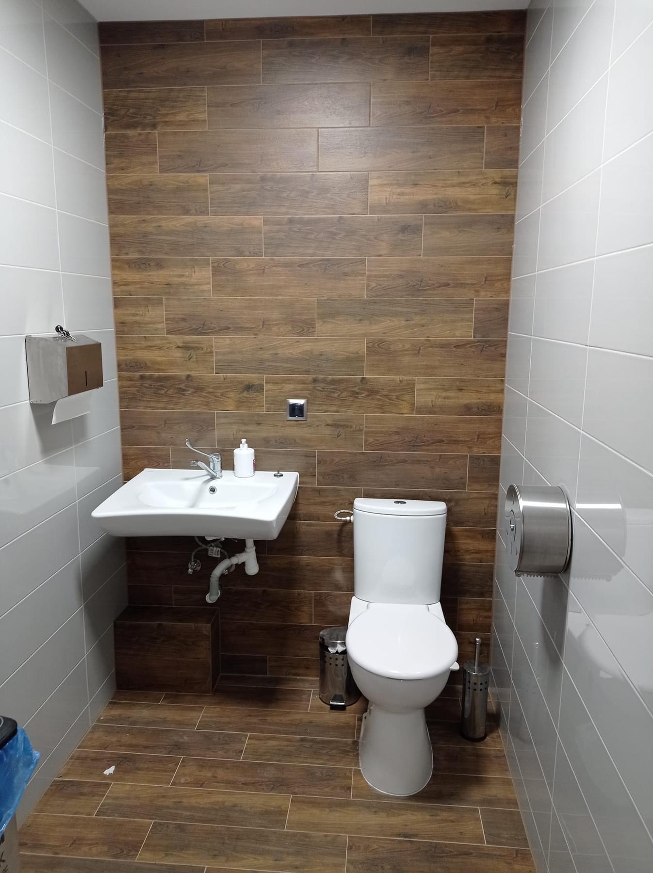 Nowoczesna łazienka z płytkami imitującymi drewno na ścianie i podłodze, biała umywalka i toaleta, metalowe akcesoria.
