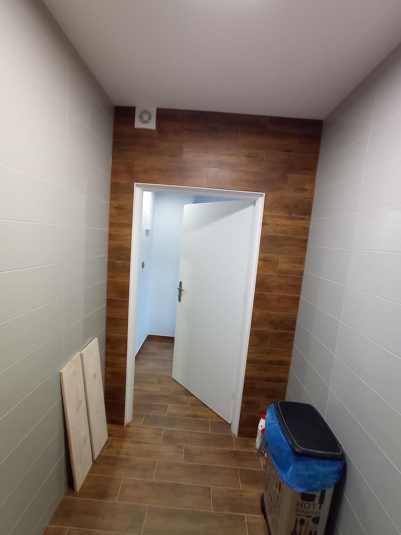 Wnętrze łazienki w trakcie prac: szare płytki na ścianach, imitacja drewna na ścianie przy wejściu i na podłodze, otwarta biała drzwi, widoczny wentylator i pozostałości po remoncie w postaci śmieci.