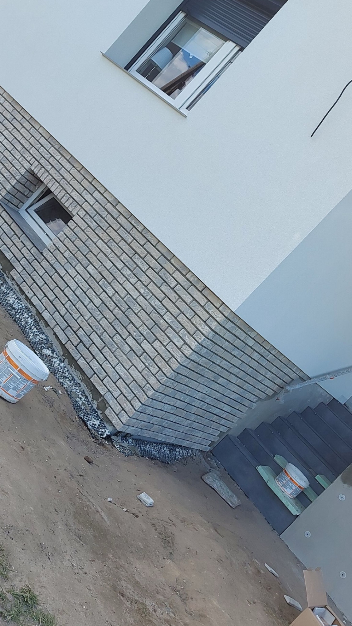 Wykończenie elewacji budynku z szarej cegły klinkierowej, widoczne okna, fragment schodów wejściowych i puszki z farbą.