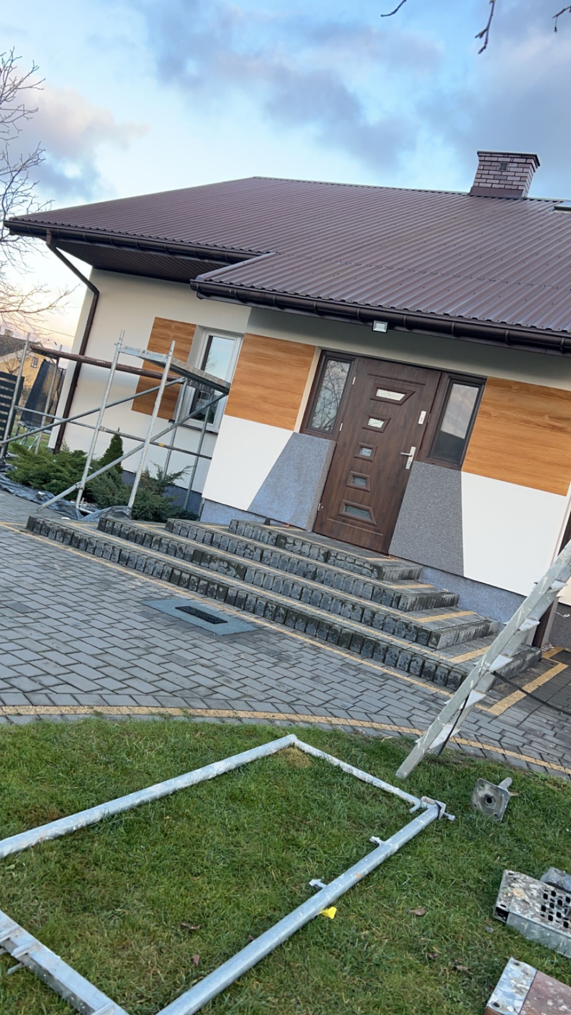 Elewacja domu jednorodzinnego z brązowym dachem i drewnopodobną okładziną, widoczne rusztowanie i schody wejściowe z kostki brukowej.