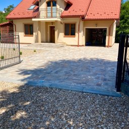 KPZ MINOR - Dwukondygnacyjny dom z garażem i podjazdem z kostki brukowej w odcieniach szarości i beżu. Otwarta brama wjazdowa. W tle zieleń drzew i błękitne niebo.