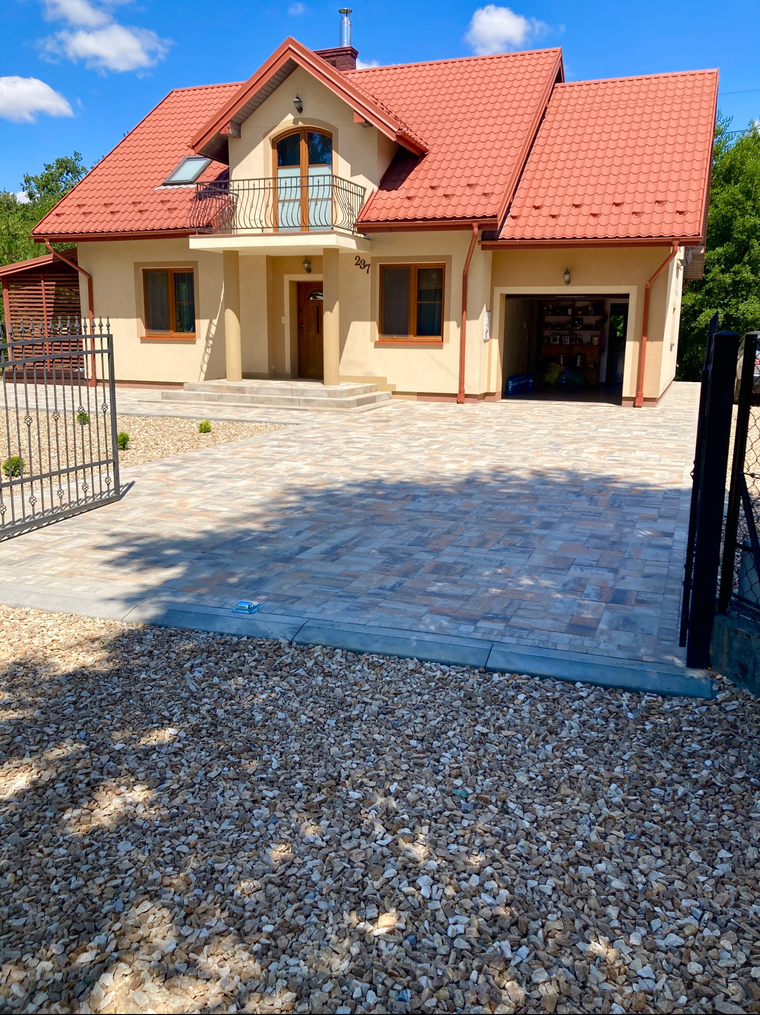 Dwukondygnacyjny dom z garażem i podjazdem z kostki brukowej w odcieniach szarości i beżu. Otwarta brama wjazdowa. W tle zieleń drzew i błękitne niebo.