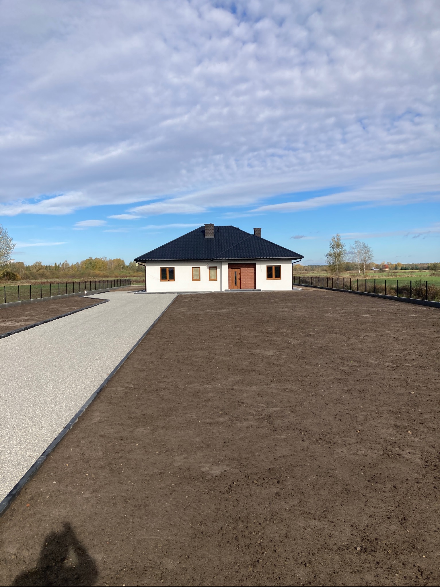 Nowoczesny dom z czarnym dachem i jasną elewacją. Podjazd z jasnego kruszywa, teren przygotowany pod trawnik. Widok na ogrodzenie i pola w tle.